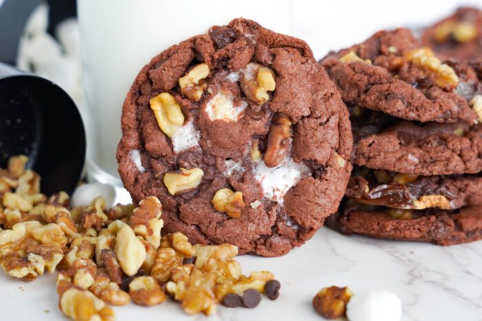 A close-up of chocolate cookies with marshmallows and walnut pieces. A glass of milk and additional walnuts are in the background.
