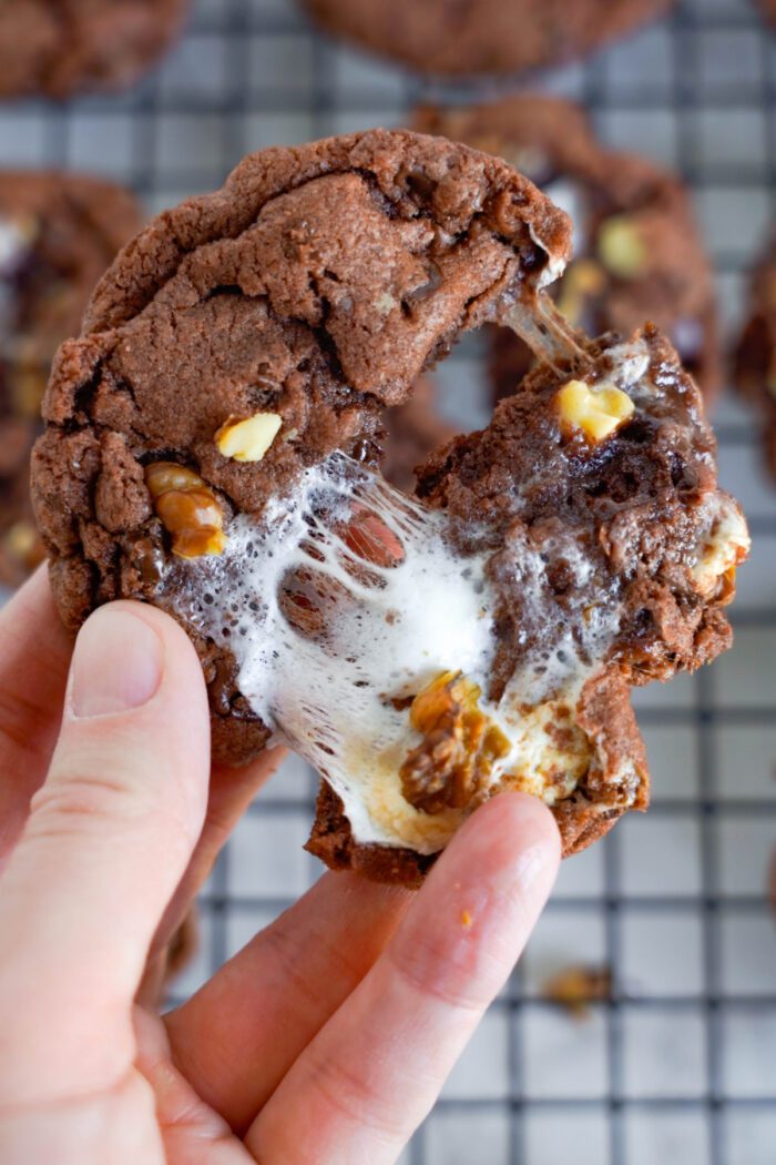 A hand holding a half-eaten chocolate cookie with melted marshmallow and nuts, highlighting its chewy texture and gooey center. Other cookies are cooling on a wire rack in the background.
