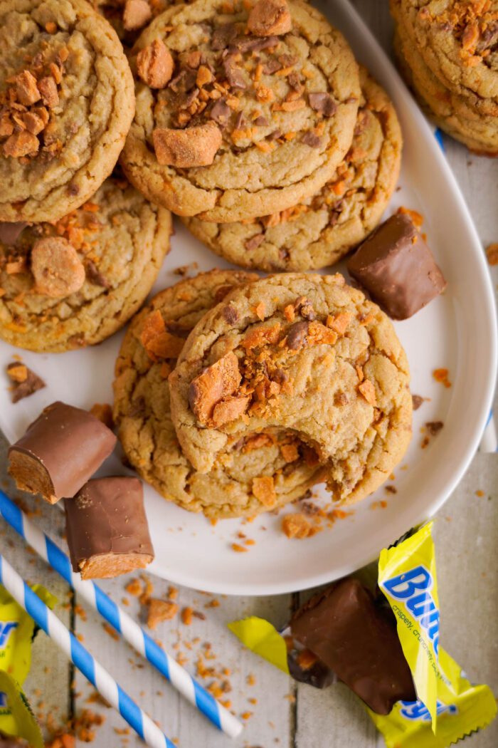 A plate of cookies topped with crumbled bits and surrounded by pieces of butterfinger and wrapped candy, with blue striped straws nearby.