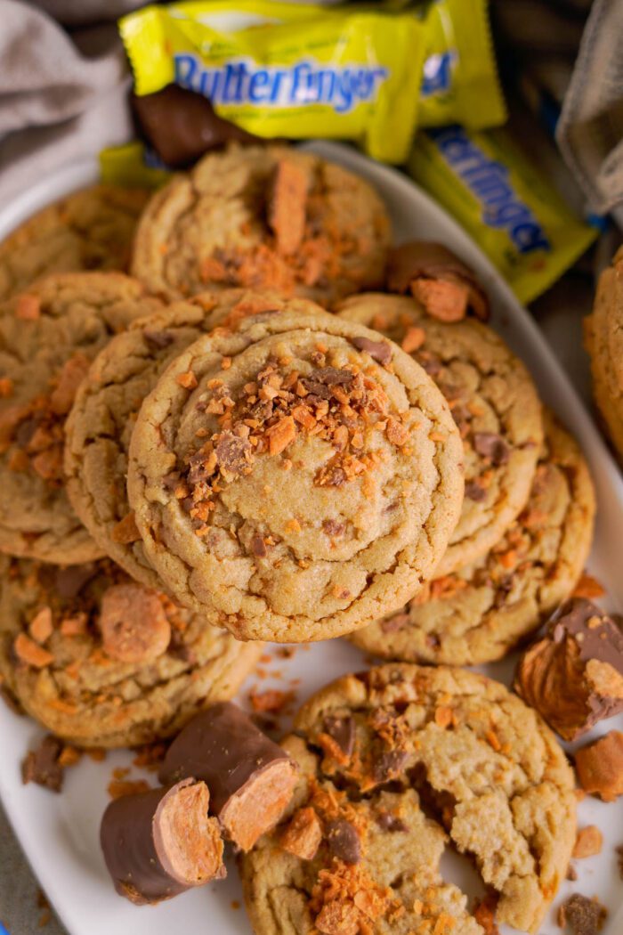 A plate of cookies topped with crumbled Butterfinger pieces, surrounded by Butterfinger candy bars, some of which are partially unwrapped.