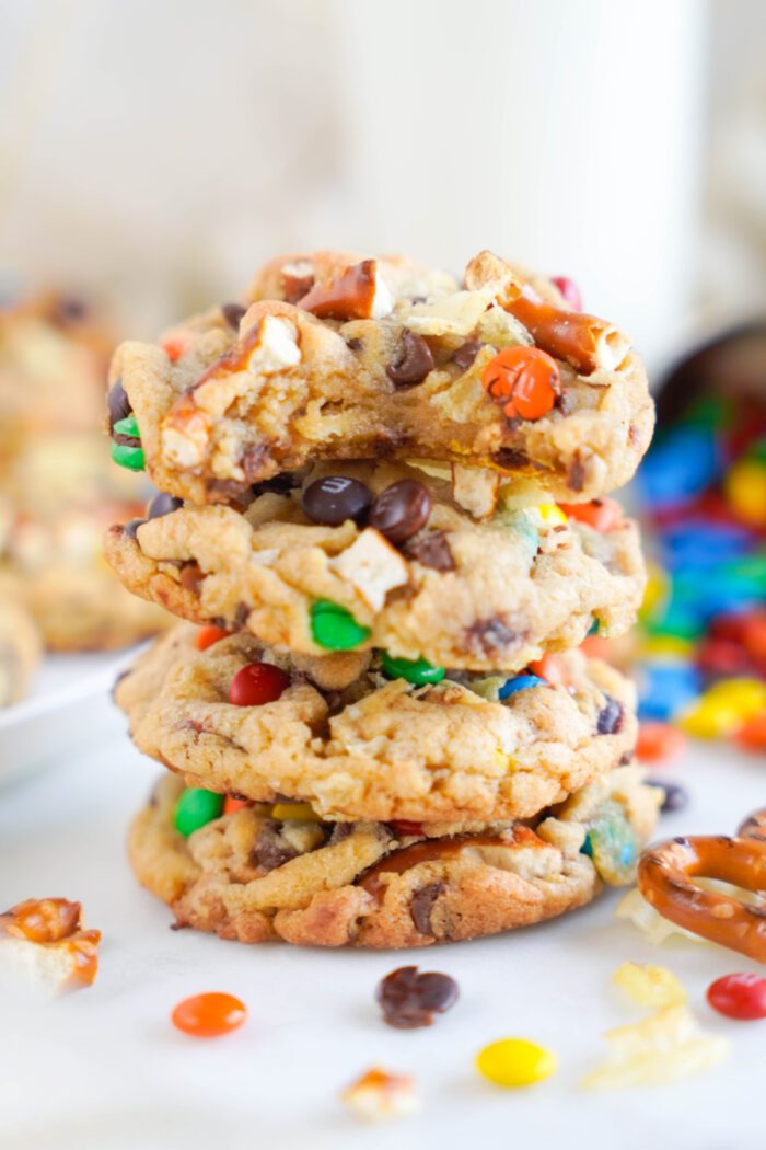 A stack of four cookies with pretzels, chocolate chips, and colorful candies on a white surface.