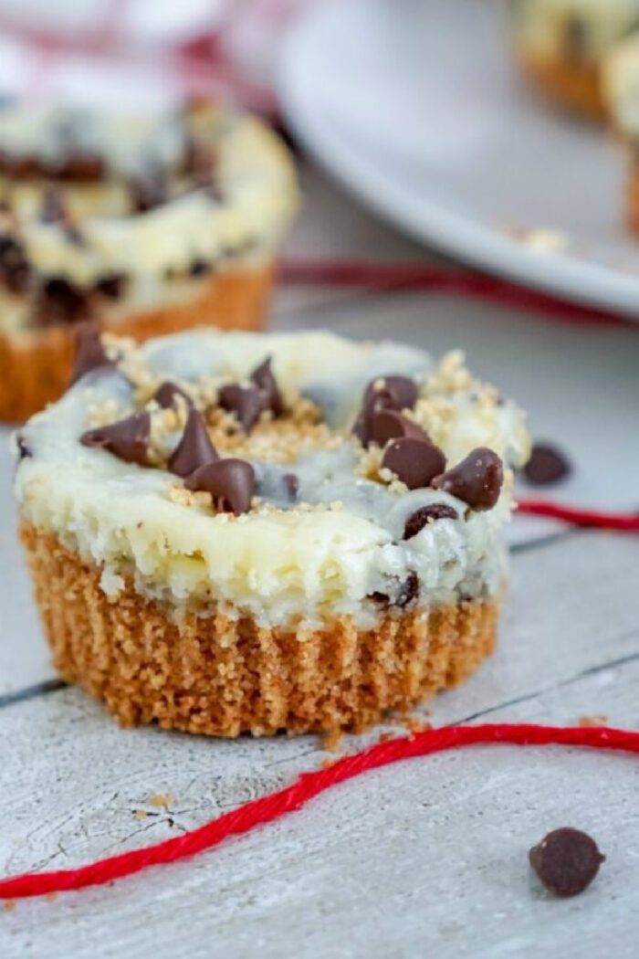 A close-up of a cheesecake cupcake topped with chocolate chips and a crumbly topping, placed on a wooden surface with a red string in the background.