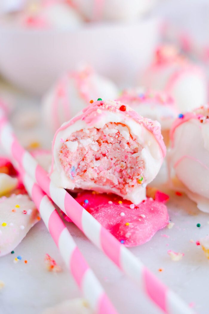 Close-up of a bitten pink and white cake ball, revealing a dense crumbly texture inside. It is decorated with sprinkles and positioned beside pink and white striped straws on a white surface.