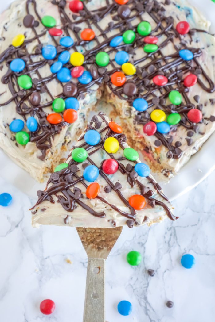 A dessert pizza topped with a creamy white frosting, colorful candy-coated chocolates, and chocolate drizzle. One slice is being lifted from the pizza on a serving utensil.