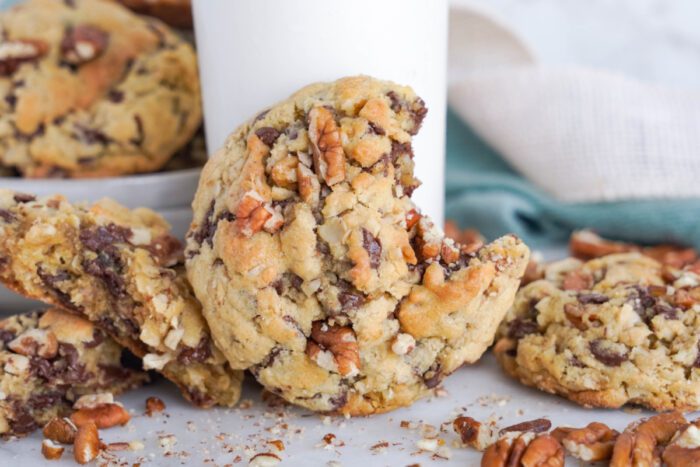 Close-up of chunky chocolate chip pecan cookies with one cookie leaning against a glass of milk. Crumbles and pecans are scattered around the cookies.