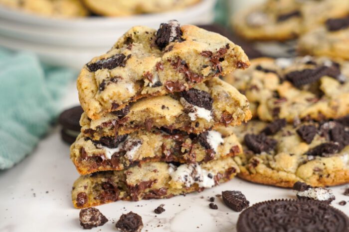 A stack of chocolate chip cookies with Oreo pieces is placed on a white surface. More cookies and a teal cloth are in the background.