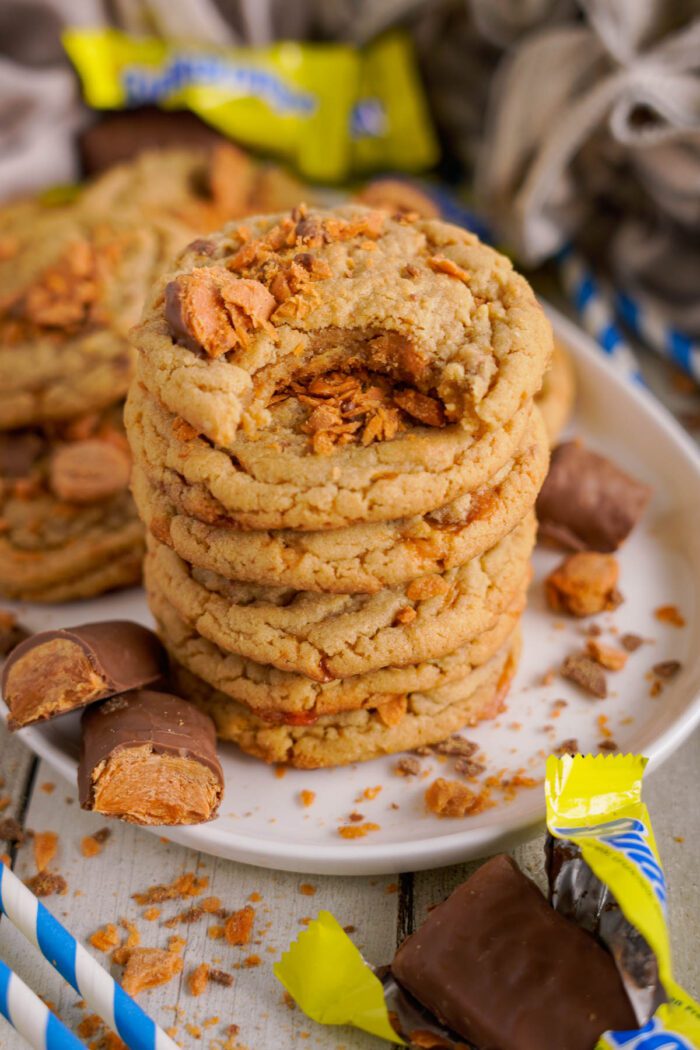 A stack of butterfinger cookies with crumbled candy pieces on top, arranged on a white plate, surrounded by partially unwrapped butterfinger candies.