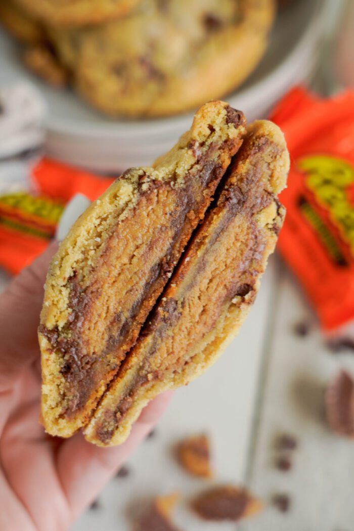 Close-up of a hand holding a halved cookie with a visible peanut butter and chocolate filling. Blurred background displays more cookies and Reese's wrapped candies.
