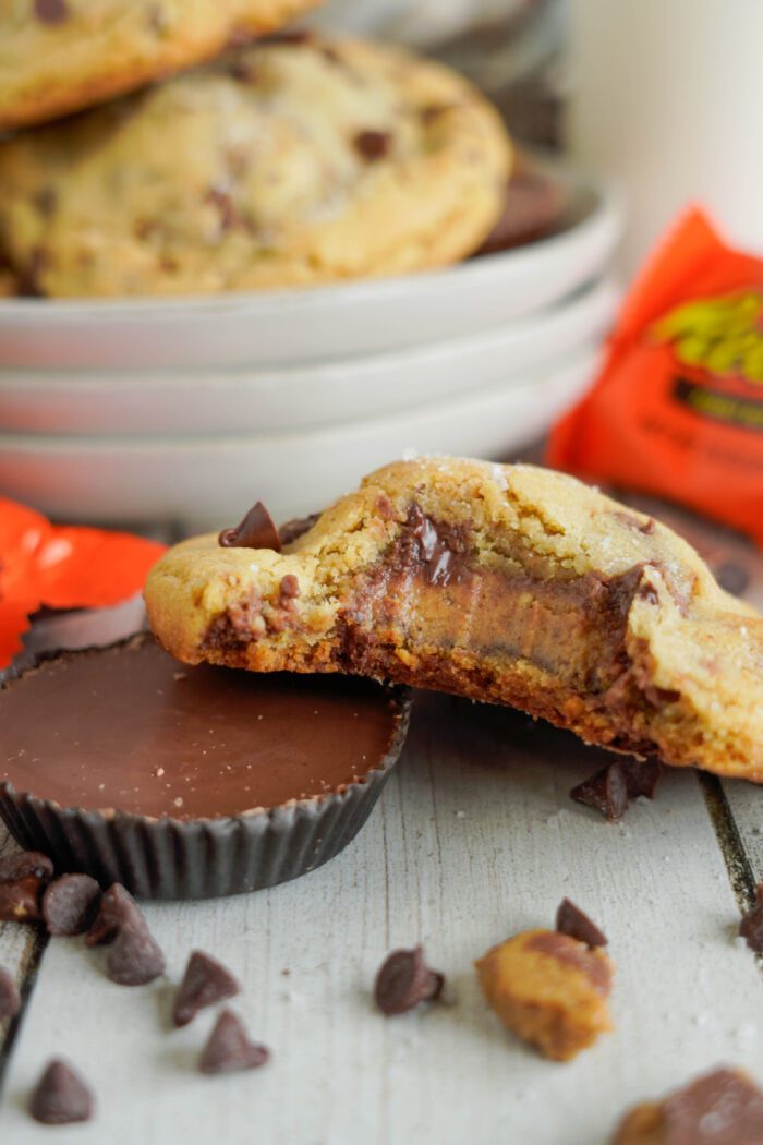 A bitten chocolate chip cookie with a peanut butter cup filling is placed in the foreground. A whole peanut butter cup and more cookies are in the background, along with scattered chocolate chips.