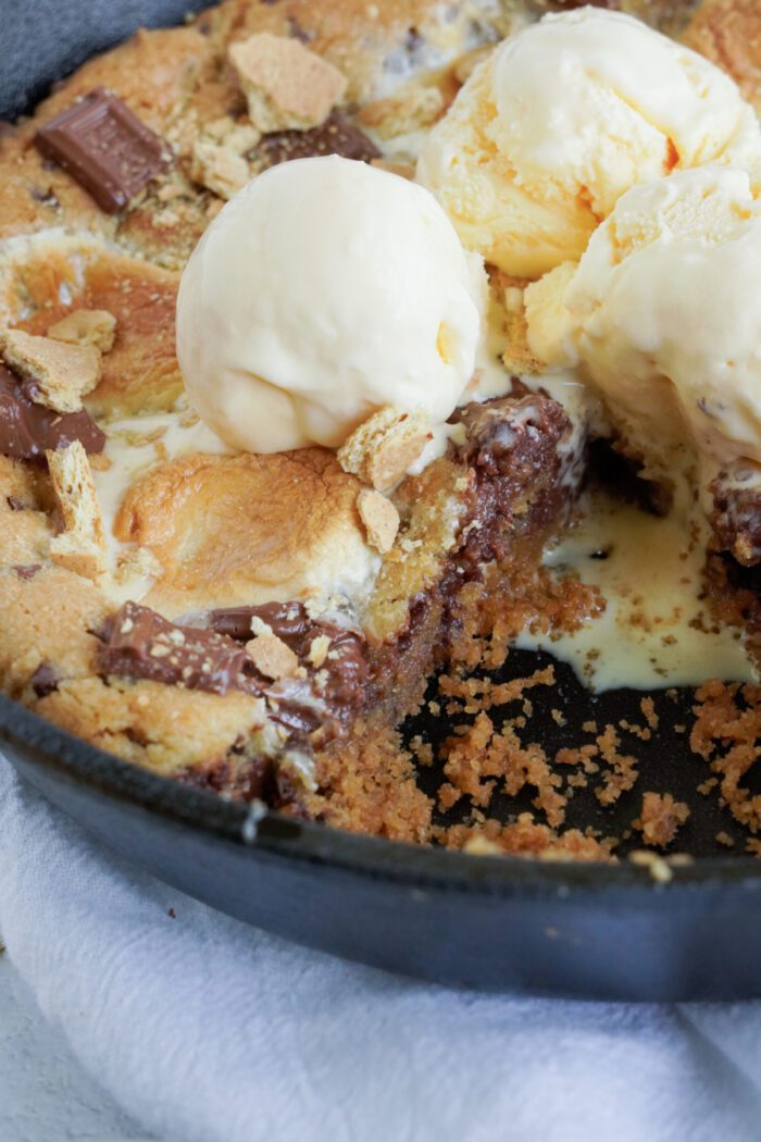 A skillet dessert with a cookie-like base, topped with marshmallows, chocolate chunks, and two scoops of vanilla ice cream is partially eaten.