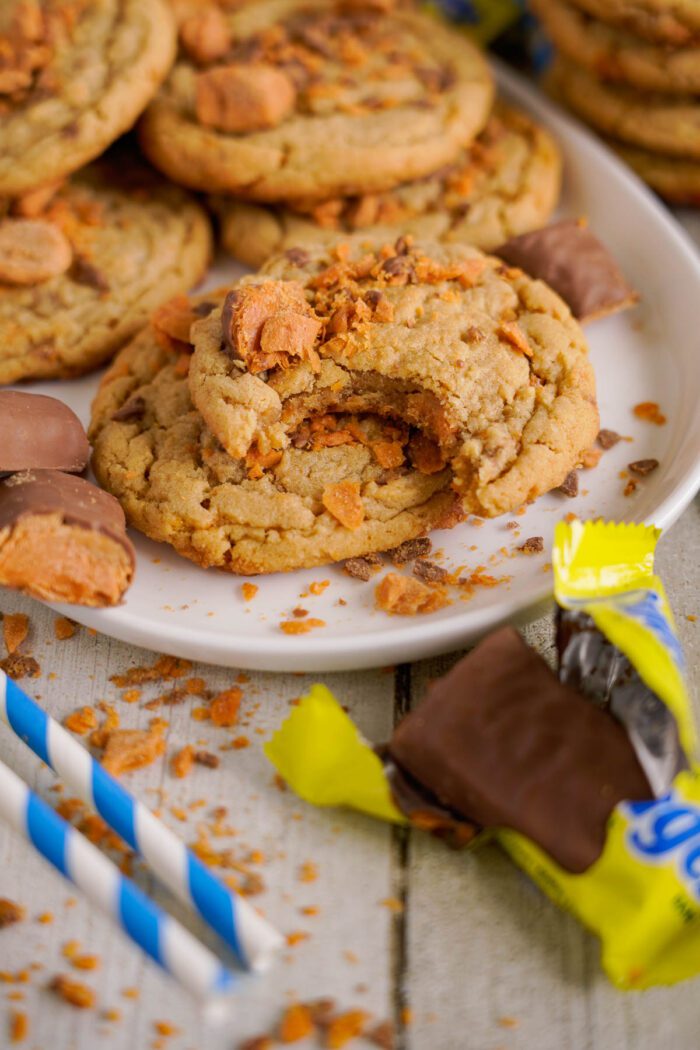 A plate of cookies with butterfinger pieces and crumbled candy toppings. One cookie has a bite taken out of it, and a few wrapped butterfinger candies are scattered around the plate.