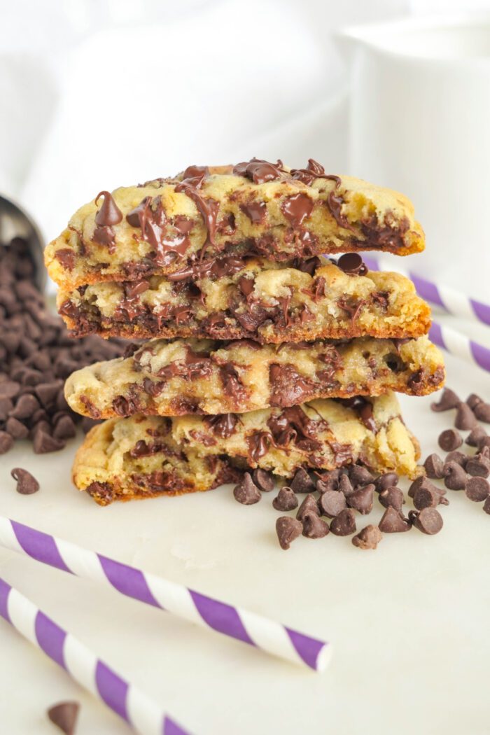 Stacked chocolate chip cookies with chocolate chips spilling around them, accompanied by purple and white striped straws on a white surface.