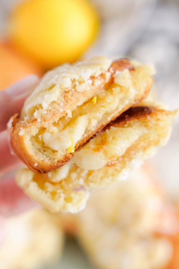 Close-up of a sliced, glazed lemon crookies revealing a crumbly interior and creamy filling. A blurred lemon and orange are in the background.