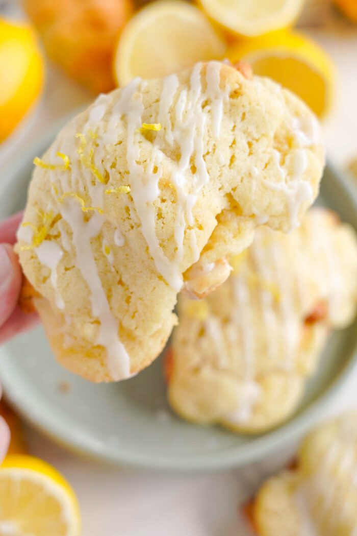 A person holds a lemon crookie drizzled with white icing. Several lemons and more cookies are visible in the background.