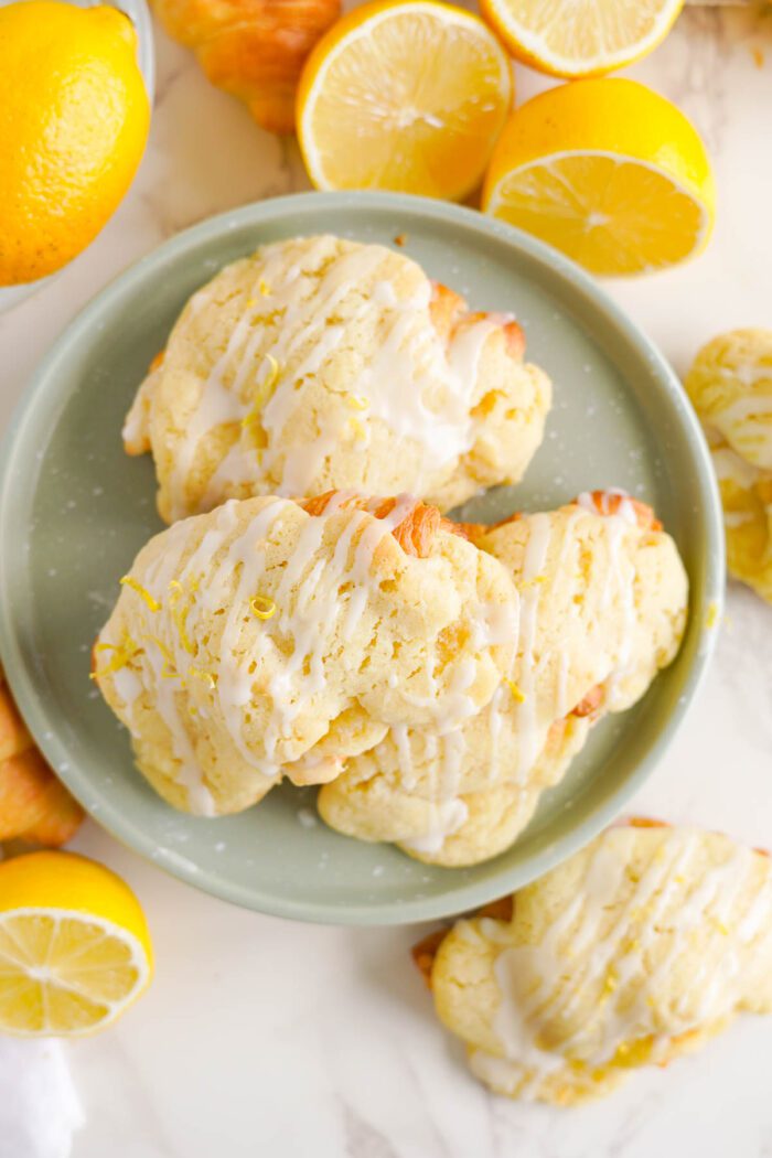 A plate of lemon crookies topped with a white glaze is surrounded by fresh lemons and lemon halves.