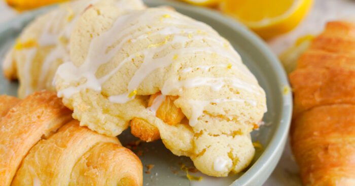 A close-up photo of croissants on a plate, with one drizzled with white icing and another plain. A sliced lemon is visible in the background.