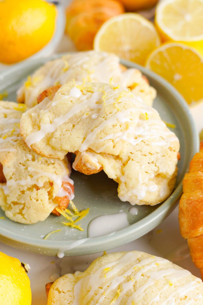A plate of glazed lemon crookies garnished with lemon zest. Sliced lemons and other pastries are in the background.