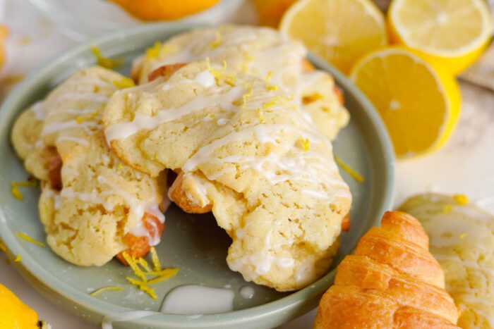 A plate holds lemon crookies drizzled with icing. Fresh lemon slices and a croissant are placed beside the plate.