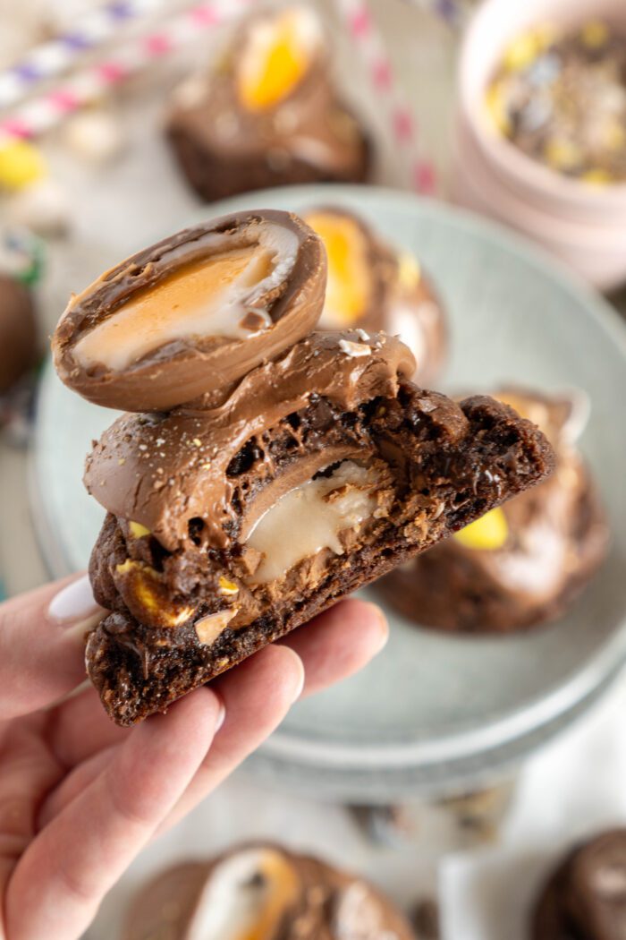 A hand holds a BIG & THICK Creme Egg Stuffed Chocolate Cookie topped with chocolate cream and a halved creme-filled egg, perfect for Easter, with more cookies and candy blurred in the background.
