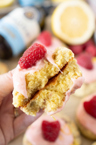 A hand holding a split cupcake topped with pink frosting and a raspberry, with more cupcakes and blurred drinks in the background.